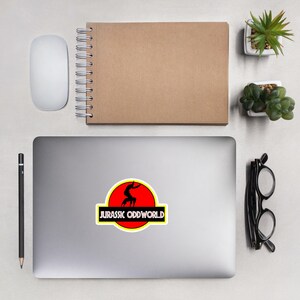 May include: A silver laptop computer with a sticker of a black silhouette of a character jumping on a red dinosaur silhouette with a yellow outline. The text "Jurassic Oddworld" is below the dinosaur.
