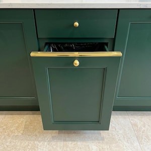 May include: A dark green kitchen cabinet with a pull-out trash drawer featuring a brass handle and a matching brass knob. The drawer is partially open, revealing a black plastic bin liner inside. The cabinet has a recessed panel design.