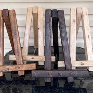 May include: A collection of small wooden easels in various shades of brown and natural wood. The easels are constructed with a simple A-frame design, perfect for displaying artwork or signage. Each easel has a horizontal support bar.