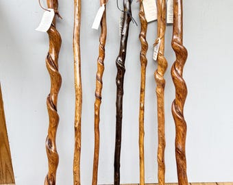 May include: Seven wooden walking sticks of varying lengths and shades of brown. Several sticks feature a natural spiral twist pattern along the shaft. Each stick includes a leather wrist strap and a small white paper tag attached near the top.
