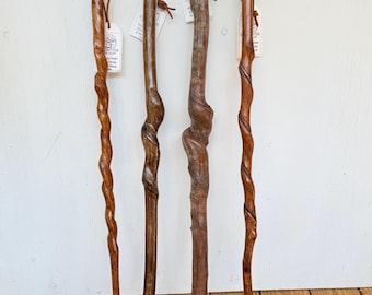 May include: Four wooden walking sticks standing upright. Each stick features a unique, natural grain pattern and texture. Two sticks have a spiral carved design, while the others retain a more organic, knotted shape. Each includes a leather strap.