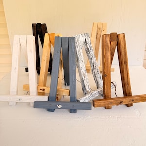 May include: A collection of six wooden tabletop easels in various finishes, including white, black, natural light wood, dark gray, distressed white, and medium brown, each featuring a hinged A-frame design with a horizontal support ledge.