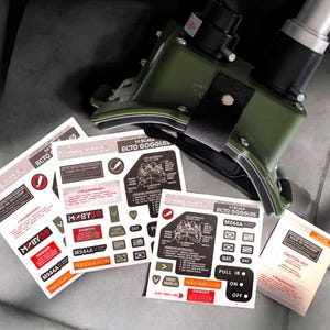 Puede incluir: Un conjunto de pegatinas para una réplica a escala 1:1 de las gafas Ecto de los Cazafantasmas. Las pegatinas están impresas en papel blanco con texto negro y rojo. Las pegatinas incluyen advertencias, instrucciones y logotipos. Las pegatinas están dispuestas sobre una superficie gris.