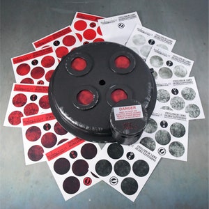 Op de afbeelding: Een zwart, rond metalen object met vier rode cirkels en een waarschuwingslabel met de tekst "GEVAAR HOGE SPANNING THY ELEKTRISCHE SCHOK GEVAAR STROOM UITSCAKELEN VOOR REPARATIE OF AANPASSING". Het object is omgeven door vellen papier met zwarte en witte cirkels en de tekst "CYCLOTRON LENS SKINS/DIFFUSERS".