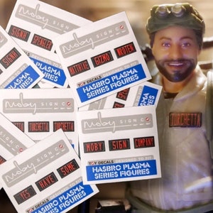 Peut inclure: Un ensemble d'autocollants blancs avec du texte noir et rouge. Les autocollants sont conçus pour s'adapter aux figurines Hasbro Plasma Series. Les autocollants présentent le texte "Moby Sign Co." et divers autres mots et expressions, notamment "Spengler", "Gizmo", "Turchetta", "Oui", "Avoir", "Certains", "Moby", "Signe" et "Compagnie".