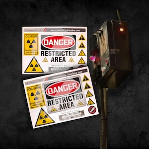 May include: Two white stickers with black and yellow warning signs. The top sticker reads "CAUTION RADIOACTIVE MATERIALS" and "PROTON PACK CHARGING UNIT" with a red and white "DANGER" sign and "RESTRICTED AREA" in black text. The bottom sticker is the same but with a different security seal number.