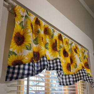 K&ouml;nnte beinhalten: Ein Gardinenvolant mit Sonnenblumenmuster. Der Vorhang zeigt gro&szlig;e gelbe Sonnenblumen mit gr&uuml;nen Bl&auml;ttern auf wei&szlig;em Grund. Der untere Rand hat eine schwarz-wei&szlig; karierte Bord&uuml;re.