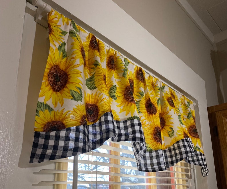 K&ouml;nnte beinhalten: Ein Gardinenvolant mit Sonnenblumenmuster. Der Vorhang zeigt gro&szlig;e gelbe Sonnenblumen mit gr&uuml;nen Bl&auml;ttern auf wei&szlig;em Grund. Der untere Rand hat eine schwarz-wei&szlig; karierte Bord&uuml;re.