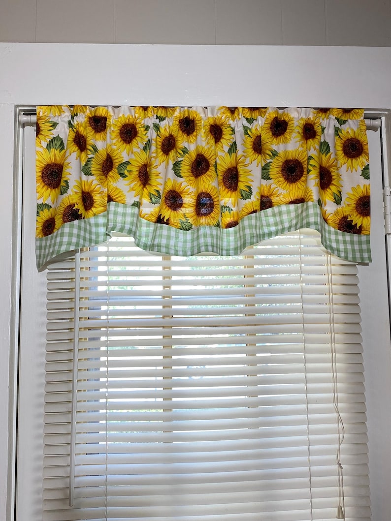 K&ouml;nnte beinhalten: Ein Fensterbehang mit einem sich wiederholenden Muster aus gelben Sonnenblumen mit braunen Zentren und gr&uuml;nen Bl&auml;ttern auf wei&szlig;em Grund. Der untere Rand hat eine gr&uuml;n-wei&szlig; karierte Bord&uuml;re. Der Fensterbehang h&auml;ngt &uuml;ber einem Fenster mit wei&szlig;en Jalousien.