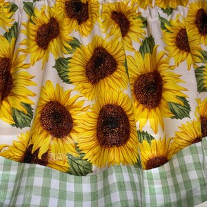 K&ouml;nnte beinhalten: Ein Fensterbehang mit einem sich wiederholenden Muster aus gro&szlig;en Sonnenblumen mit gelben Bl&uuml;tenbl&auml;ttern und braunen Zentren auf wei&szlig;em Hintergrund. Der untere Rand hat eine gr&uuml;n-wei&szlig; karierte Bord&uuml;re. Der Stoff ist oben gerafft.