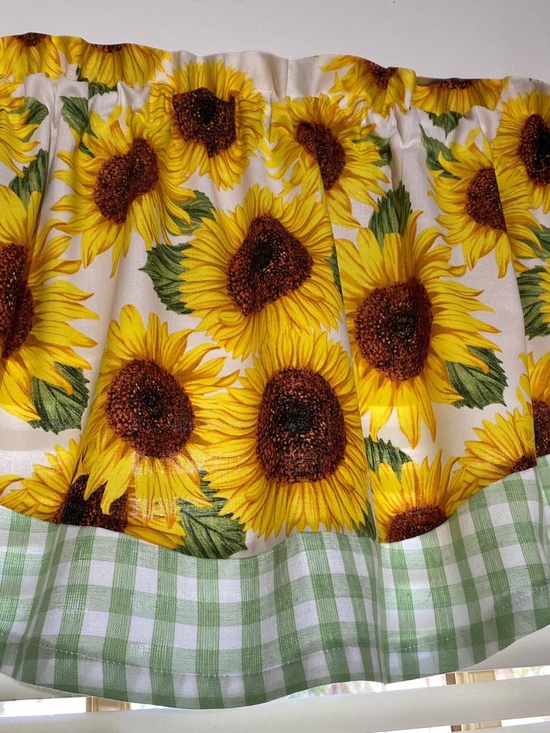 K&ouml;nnte beinhalten: Ein Fensterbehang mit einem sich wiederholenden Muster aus gro&szlig;en Sonnenblumen mit gelben Bl&uuml;tenbl&auml;ttern und braunen Zentren auf wei&szlig;em Hintergrund. Der untere Rand hat eine gr&uuml;n-wei&szlig; karierte Bord&uuml;re. Der Stoff ist oben gerafft.