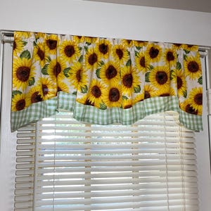 K&ouml;nnte beinhalten: Ein Fensterbehang mit einem sich wiederholenden Muster aus gelben Sonnenblumen mit braunen Zentren und gr&uuml;nen Bl&auml;ttern auf wei&szlig;em Hintergrund. Der untere Rand hat eine gr&uuml;n-wei&szlig; karierte Bord&uuml;re. &Uuml;ber wei&szlig;en Jalousien.
