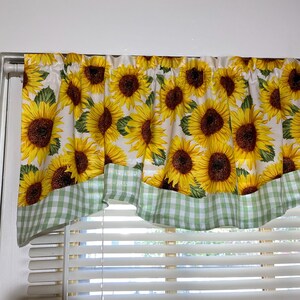 K&ouml;nnte beinhalten: Ein Fensterbehang mit einem sich wiederholenden Muster aus gelben Sonnenblumen mit braunen Zentren und gr&uuml;nen Bl&auml;ttern auf wei&szlig;em Hintergrund. Der untere Rand ist mit einem gr&uuml;n-wei&szlig; karierten Stoff verziert. Der Behang h&auml;ngt &uuml;ber einem Fenster mit wei&szlig;en Jalousien.