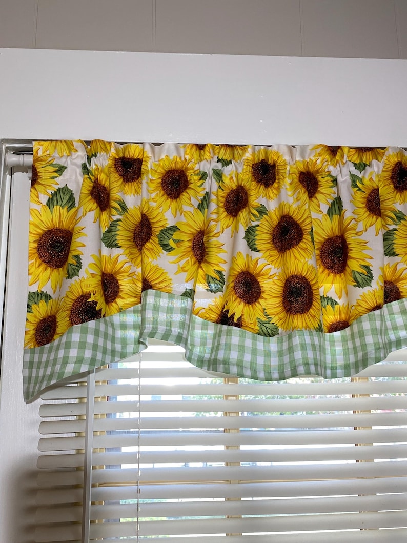 K&ouml;nnte beinhalten: Ein Fensterbehang mit einem sich wiederholenden Muster aus gelben Sonnenblumen mit braunen Zentren und gr&uuml;nen Bl&auml;ttern auf wei&szlig;em Hintergrund. Der untere Rand ist mit einem gr&uuml;n-wei&szlig; karierten Stoff verziert. Der Behang h&auml;ngt &uuml;ber einem Fenster mit wei&szlig;en Jalousien.