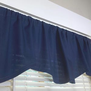 May include: A navy blue valance curtain with a scalloped bottom edge is hung on a white curtain rod above a window with white blinds. The curtain is made of a textured fabric and is gathered along the rod.