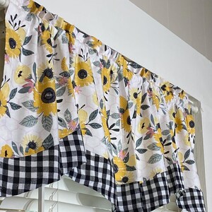 K&ouml;nnte beinhalten: Ein Fensterbehang mit wei&szlig;em Hintergrund, verziert mit gelben Sonnenblumen, rosa Akzenten und grauen Bl&auml;ttern. Der untere Rand hat ein schwarz-wei&szlig;es Karomuster. Der Fensterbehang h&auml;ngt &uuml;ber einem Fenster.