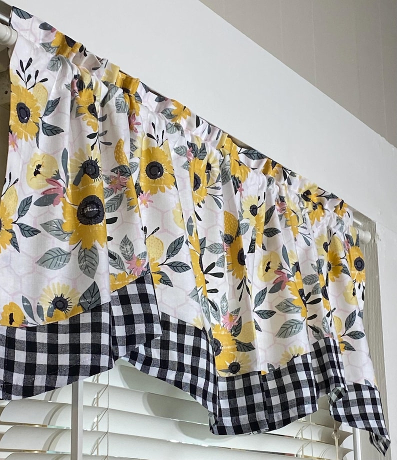 K&ouml;nnte beinhalten: Ein Fensterbehang mit wei&szlig;em Hintergrund, verziert mit gelben Sonnenblumen, rosa Akzenten und grauen Bl&auml;ttern. Der untere Rand hat ein schwarz-wei&szlig;es Karomuster. Der Fensterbehang h&auml;ngt &uuml;ber einem Fenster.