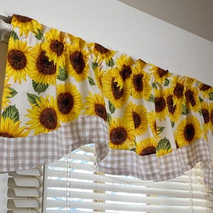 K&ouml;nnte beinhalten: Ein Fensterbehang mit einem sich wiederholenden Muster aus gelben Sonnenblumen mit braunen Zentren und gr&uuml;nen Bl&auml;ttern auf wei&szlig;em Grund. Der untere Rand hat eine braun-wei&szlig; karierte Bord&uuml;re. Der Behang h&auml;ngt &uuml;ber einem Fenster.