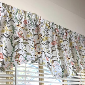 May include: A floral valance with a white background and a pattern of colorful wildflowers. The flowers include red, pink, yellow, and white blooms with green foliage. The valance is hanging above a window with white blinds.
