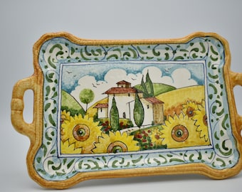 Könnte beinhalten: Rechteckige Servierplatte aus Keramik mit handgemaltem italienischem Landschaftsmotiv. Das Tablett hat einen gelb-blauen Rand mit Griffen. Das zentrale Bild zeigt ein Haus, Sonnenblumen und sanfte Hügel unter blauem Himmel.