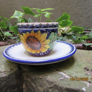 May include: A white ceramic teacup and saucer with a blue rim and a sunflower design. The cup features a blue and yellow sunflower with green leaves.