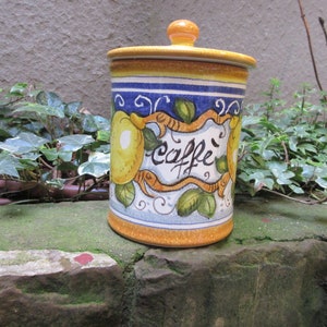 May include: A yellow, blue, and white ceramic canister with a lemon design and the word "caffe" written on it. The canister has a lid and is sitting on a stone surface.