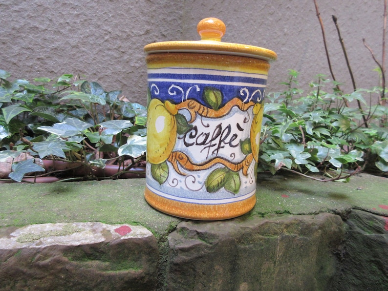 May include: A yellow, blue, and white ceramic canister with a lemon design and the word "caffe" written on it. The canister has a lid and is sitting on a stone surface.