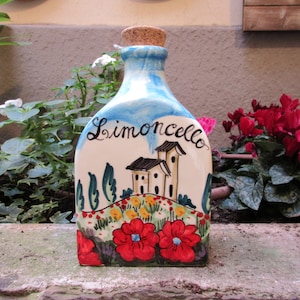 May include: A hand-painted ceramic bottle with a cork stopper. The bottle is decorated with a blue and white sky, green trees, a white house with red tile roof, and red flowers. The bottle says "Limoncello" in black cursive writing.
