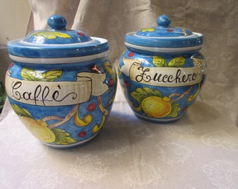 Op de afbeelding: Twee blauwe en witte keramische potten met deksels. De potten zijn versierd met citroenen, bladeren en wijnstokken. Eén pot zegt "Caffe'" en de andere zegt "Zucchero".