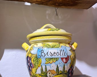 May include: A yellow ceramic jar with a lid and two side handles, featuring a hand-painted landscape scene with a house, cypress trees, grapes, and apples. A blue banner across the front displays the word Biscotti in black cursive script.