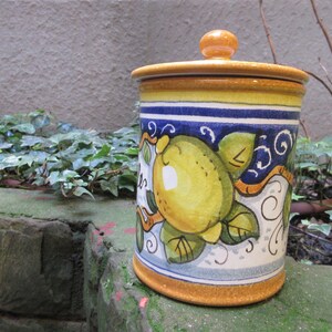 May include: A ceramic canister with a yellow, blue, and white lemon design. The canister has a lid with a knob on top. The canister is sitting on a stone surface.