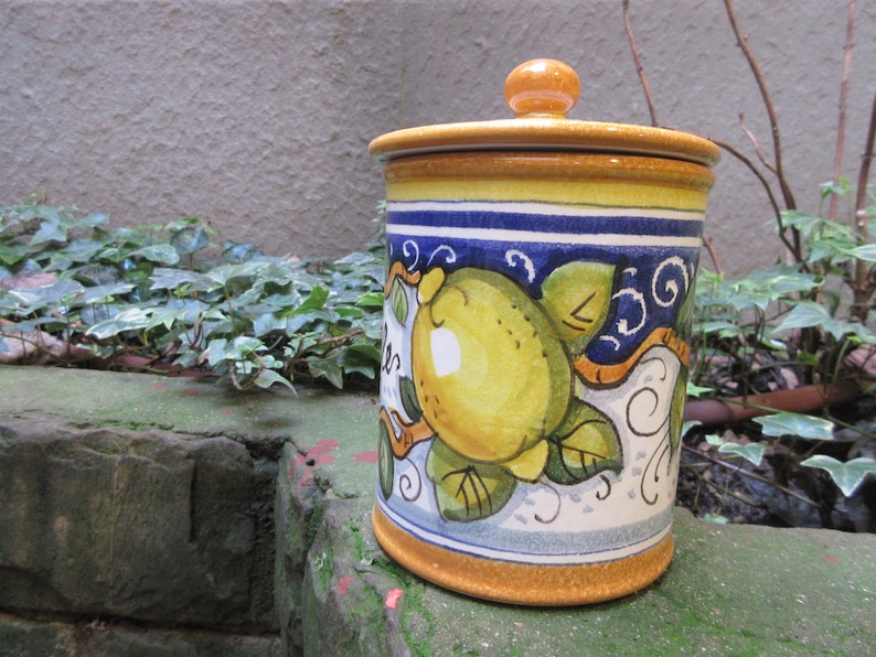 May include: A ceramic canister with a yellow, blue, and white lemon design. The canister has a lid with a knob on top. The canister is sitting on a stone surface.