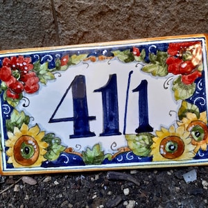 Può includere: Un cartello di numero civico in ceramica bianca con numeri blu "41/1" circondato da un disegno floreale di fiori rossi, foglie verdi e girasoli gialli su sfondo blu.