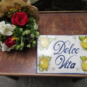 Może przedstawiać: Drewniany stół z bukietem kwiatów owiniętym w brązowy papier i białą ceramiczną płytką z napisem "Dolce Vita" i namalowanymi cytrynami.