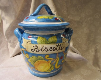 May include: A blue and white ceramic cookie jar with a painted design of lemons and leaves. The jar has a lid with a handle and says "Biscotti" on the front.