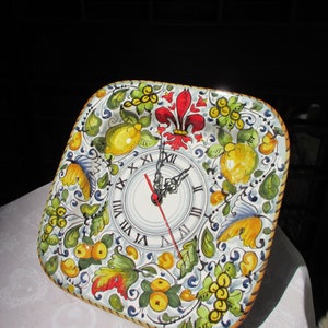 Peut inclure: Horloge murale carrée en céramique avec un motif peint à la main. Le cadran de l'horloge comporte des chiffres romains et des aiguilles noires. La bordure est ornée de citrons, de feuilles et d'une fleur de lys rouge. L'horloge mesure environ 25 cm de côté.