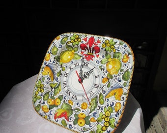 May include: Square ceramic wall clock with a hand-painted design. The clock face features Roman numerals and black clock hands. The border is decorated with lemons, leaves, and a red fleur-de-lis. The clock measures approximately 10 inches square.