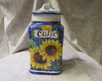 Op de afbeelding: Een blauwe en witte keramische koffiebus met een zonnebloemdessin en het woord "Caffe'" op de voorkant.