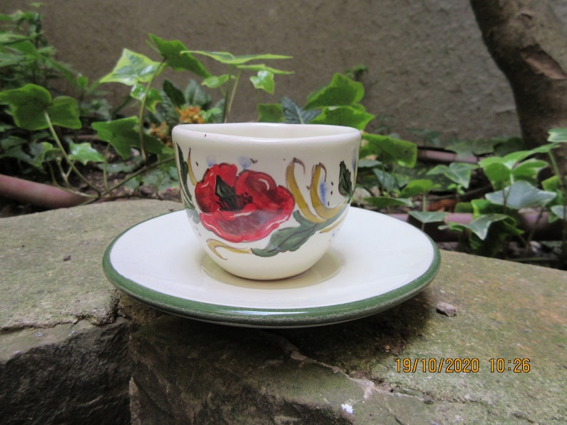 May include: A white ceramic teacup and saucer with a green rim. The cup and saucer are decorated with a red poppy flower and green leaves.