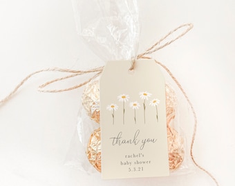 Op de afbeelding: Een beige bedankkaartje met een wit madeliefje-ontwerp en de tekst "bedankt, rachel's babyshower, 5.3.21". Het kaartje is vastgebonden aan een doorzichtige plastic zak gevuld met in gouden folie verpakte chocolaatjes.
