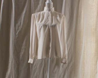 May include: A cream-colored blouse with a high collar and long sleeves. The blouse is buttoned down the front and has a simple, elegant design. The sleeves have buttoned cuffs. The blouse is displayed on a white mannequin.
