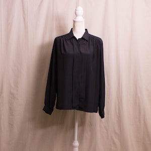 May include: A black long-sleeve blouse with a collared neckline and button-down front. The blouse is displayed on a white mannequin. The fabric appears to be a smooth, solid material.