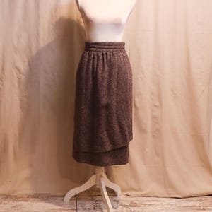 May include: A brown and grey tweed midi skirt featuring a high-waisted design with a thick, ribbed waistband and a layered, wrap-style front panel. The skirt is displayed on a white mannequin, showcasing its textured fabric and knee-length cut.