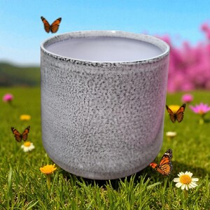 May include: A cylindrical, gray ceramic container with a textured surface. The container has a light-colored interior and a dark rim. Orange butterflies and wildflowers are in the background.