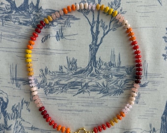 May include: A colorful beaded necklace with a gradient of colors, including red, orange, yellow, and pink. The necklace has a gold-colored clasp and is displayed on a blue and white patterned fabric.