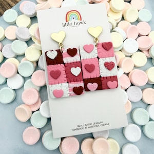 Op de afbeelding: Oorbellen met gouden hartvormige toppen en rechthoekige hangers. De hangers zijn in patchwork-stijl, met vierkanten in wit, roze en bordeauxrood, en hartaccenten. De kaart toont "little hawk" en "Small Batch Jewelry, Handmade in Manitoba, Canada."