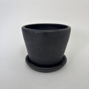 May include: A small, black ceramic plant pot with a matching saucer. The pot has a slightly tapered shape and a matte finish. The saucer is round and fits snugly beneath the pot. Suitable for small plants or succulents.