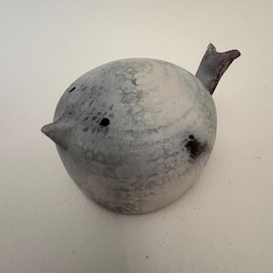May include: A ceramic bird ornament with a rounded body and a small, pointed beak. The bird is mainly grey with darker grey and black markings. It has a small, dark tail. The figurine is a decorative item.