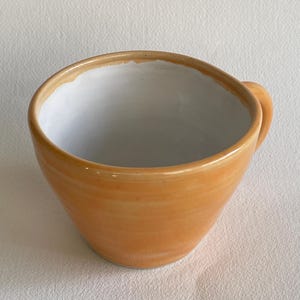 May include: A ceramic mug with a white interior and a light orange exterior. The mug has a rounded shape and a small handle.