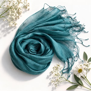 Peut inclure: Une écharpe bleu sarcelle avec une texture tissée visible et des détails de franges délicates sur les bords. Le tissu est léger et semi-transparent, disposé en un motif circulaire plié qui souligne la couleur bleu-vert profond du matériau.